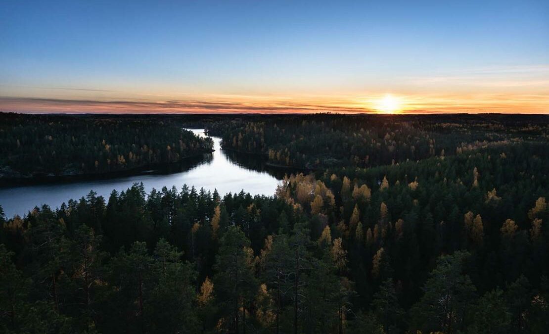 Ilmakuva Repoveden kansallispuistosta illan hämärässä. Edessä tummasävyistä metsää, keskellä vettä ja taivas on sinioranssi.