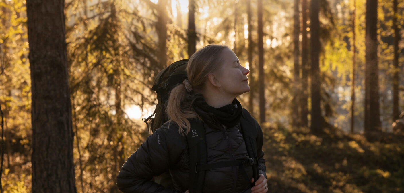 Nainen retkivarusteissa seisoo metsässä kasvot kohotettuina ja silmät kiinni. Viisto auringonvalo kultaa taustan puut.