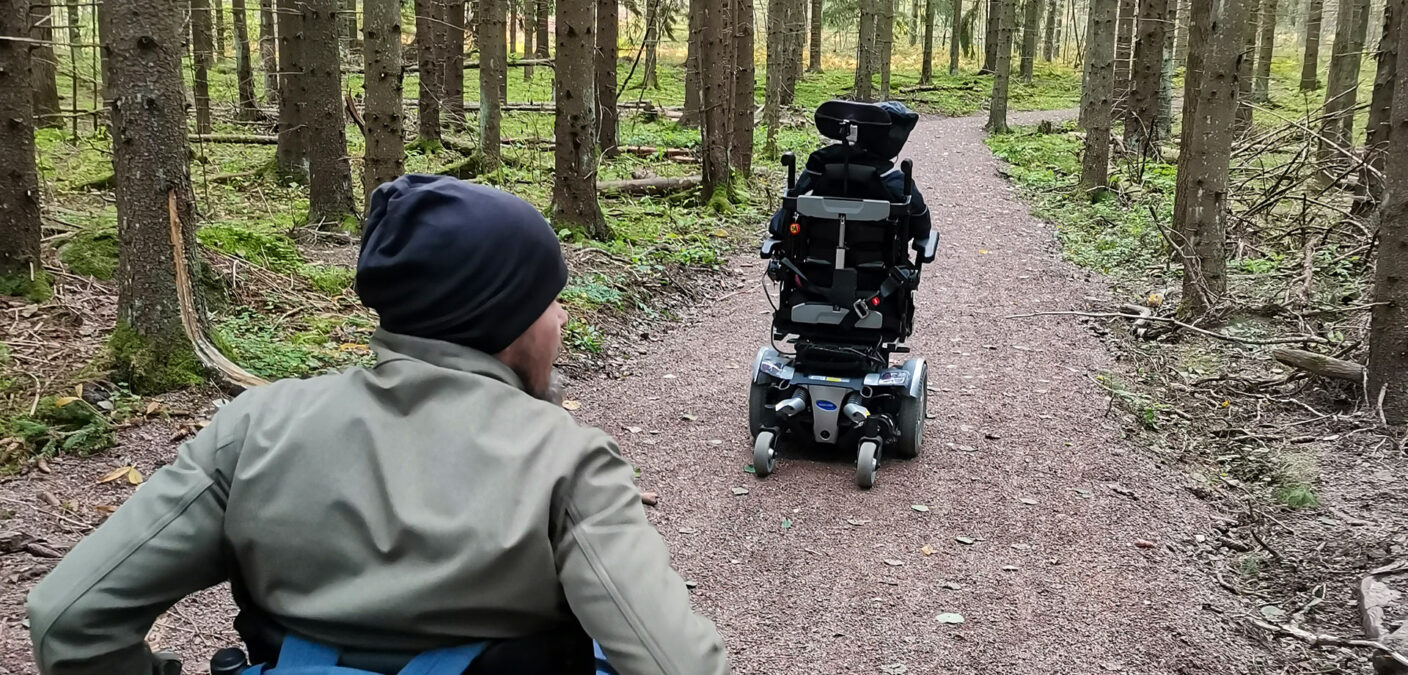 Kaksi henkilöä kulkee pyörätuoleilla Kumijoen ulkoilureitillä metsässä.
