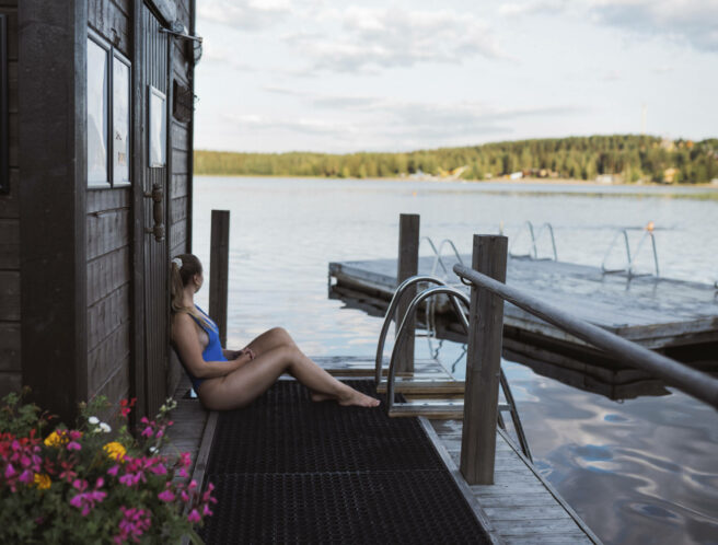 Nainen istuu rantasaunan laiturilla ja katselee järvelle päin.